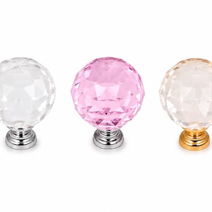 May include: Three spherical cabinet knobs with faceted surfaces. The left knob is clear with a silver-tone base, the center knob is pink with a silver-tone base, and the right knob is clear with a gold-tone base. Each knob features a round, multi-faceted top.