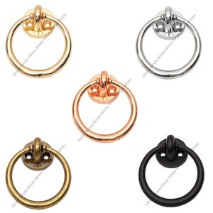 Op de afbeelding: Vijf metalen ringgrepen in goud, zilver, koper, antiek messing en zwart. Elke greep heeft een cirkelvormige ring bevestigd aan een ronde basis met twee schroefgaten. Deze decoratieve hardware is geschikt voor kasten en laden.