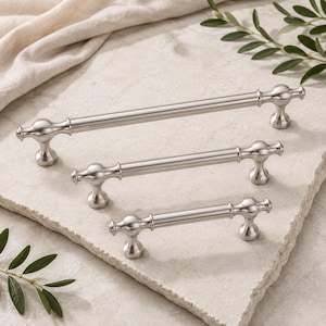 May include: Three brushed nickel cabinet pulls of varying lengths arranged diagonally. Each handle features a smooth cylindrical bar with rounded, flared ends and a pedestal base, showcasing a consistent metallic finish and uniform design.