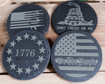 Op de afbeelding: Vier ronde zwarte leisteen onderzetters met verschillende patriottische ontwerpen. De onderzetters tonen een Amerikaanse vlag met wapens, een slang met de tekst "Don't Tread On Me", een cirkel van sterren met het jaar "1776" en een Amerikaanse vlag met de tekst "We The People".