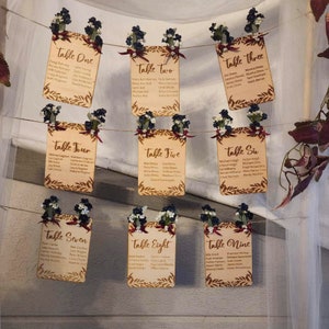 May include: Nine rectangular wooden table seating cards hanging on twine. Each card features laser-engraved text from Table One to Table Nine, guest names, and decorative leaf borders. Small dark blue and white floral sprigs are attached with ribbons.