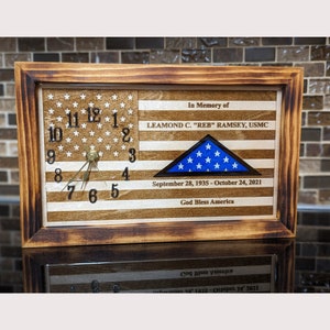 May include: A wooden clock with a US flag design, featuring a blue triangle flag with white stars. The clock face is surrounded by a wooden frame and includes the text "In Memory of LEAMOND C. "REB" RAMSEY, USMC September 28, 1935-October 24, 2021 God Bless America".