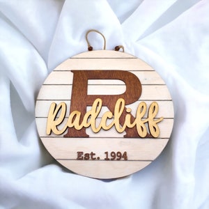 May include: A round wooden sign with a white background and brown woodgrain stripes. The sign features a large brown letter "R" and the name "Radcliff" in a cursive font. The text "Est. 1994" is written below the name.