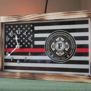 May include: A wooden framed clock with a black and white American flag design. The clock face has a red stripe across the middle and a black and white circle with the text "BURLINGTON COUNTY PROFESSIONAL FIRE FIGHTERS ASSN. INTERNATIONAL ASSOCIATION OF FIRE FIGHTERS AFL-CIO CLC LOCAL 3091".