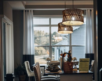 May include: A modern kitchen with a white island and bar stools. The island has a white worktop and a stainless steel sink. There are three bar stools with brown upholstery and black metal frames. The kitchen has a large window with a view of trees and a house in the distance. The kitchen also has a light fixture with three woven shades hanging over the island.