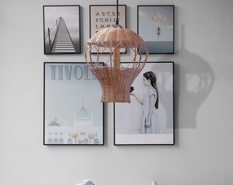May include: A wicker hot air balloon pendant light fixture hangs from the ceiling in a room with white walls and a light grey couch. The light fixture is brown and has a black cord. The room is decorated with framed posters and a white step stool.