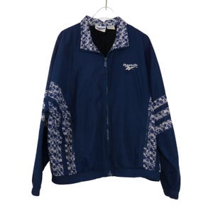 Op de afbeelding: Marineblau Reebok trainingsjack met een volledige ritssluiting en witte bloempatronen op de kraag en mouwen. Het Reebok-logo is op de borst geborduurd. De jas hangt aan een metalen hanger.