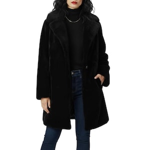 Puede incluir: Un abrigo negro de piel sintética con solapas y dos bolsillos delanteros. El abrigo tiene cierre de botón y una silueta larga y recta. La modelo lleva un cuello alto negro y vaqueros azules.