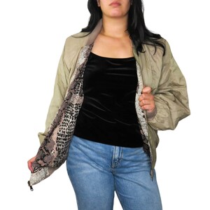 May include: A tan bomber jacket with a snake print lining. The jacket is open, revealing the patterned interior. The model is wearing a black top and blue jeans. The jacket has a zipper closure.