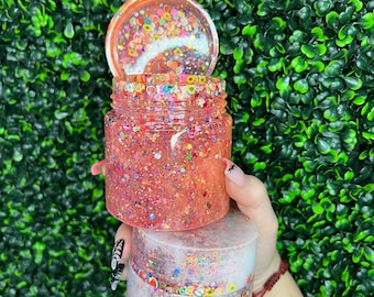 May include: Two clear glass jars with lids filled with colorful glitter and confetti. The jars are stacked on top of each other and are being held by a hand.