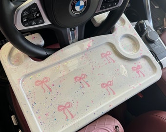 May include: White car tray with pink bows and glitter. The tray is sitting in a car with a pink purse on the seat.