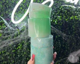 May include: Three stacked, translucent resin jars in shades of green and blue, with glitter accents. The jars are cylindrical with screw-on lids. The background features a neon sign and a green wall.