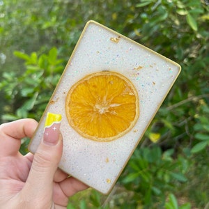 Op de afbeelding: Een vierkante witte onderzetter met gouden glitter en een gedroogde sinaasappelschijf in het midden. De onderzetter heeft een gouden rand.