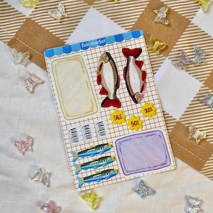 Könnte beinhalten: Ein Stickerbogen mit dem Thema "Fischmarkt". Er zeigt Illustrationen von Fischen, leere Felder für Notizen und Verkaufsetiketten. Der Bogen ist mit gelben, rosa und transparenten schmetterlingsförmigen Verzierungen dekoriert.
