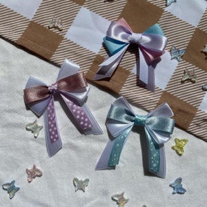 May include: Three pastel-colored satin bows with white and star-shaped patterns. The bows are arranged on a white fabric background with iridescent butterfly embellishments.