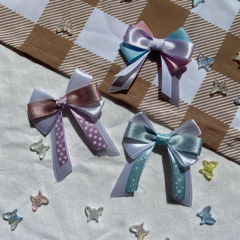 May include: Three pastel-colored satin bows with white and star-shaped patterns. The bows are arranged on a white fabric background with iridescent butterfly embellishments.