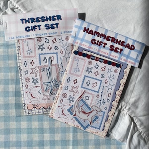 Könnte beinhalten: Zwei Geschenksets mit Hai-Motiven. Eines ist mit "THRESHER GIFT SET" und das andere mit "HAMMERHEAD GIFT SET" beschriftet. Jedes Set enthält eine Postkarte, ein Stickerbogen und Aufkleber. Die Designs zeigen Haie, Sterne und stempelförmige Rahmen in Rot, Blau und Weiß.