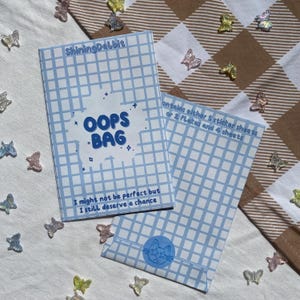 Könnte beinhalten: Eine blaue und weiße karierte Papiertüte mit dem Text "OOPS BAG" in einem blauen Kreis. Die Tüte trägt den Text "Ich bin vielleicht nicht perfekt, aber ich verdiene trotzdem eine Chance" und "enthält entweder 5 Stickerbögen oder 2 Flocken und 4 Bögen".