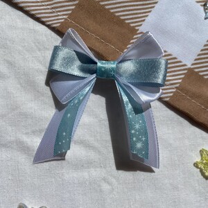 May include: A light blue and white bow with a snowflake pattern. The bow is made of satin ribbon and is tied in a simple knot.