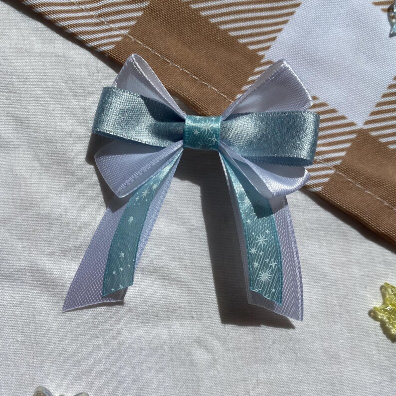 May include: A light blue and white bow with a snowflake pattern. The bow is made of satin ribbon and is tied in a simple knot.