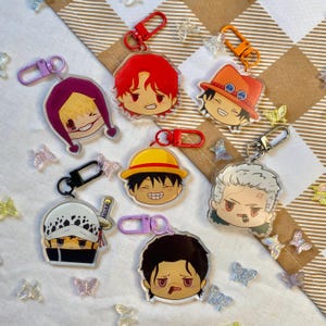 Könnte beinhalten: Eine Sammlung von Schlüsselanhängern mit Anime-Charakteren. Jeder Schlüsselanhänger zeigt das Gesicht eines anderen Charakters mit leuchtenden Farben und detaillierten Gesichtsausdrücken. Die Schlüsselanhänger haben Metallverschlüsse in verschiedenen Farben.