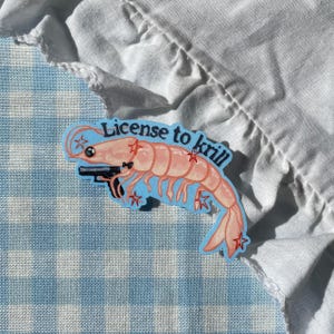May include: A sticker featuring a cartoon shrimp holding a gun, with the text "License to krill". The shrimp is pink with red star accents on a light blue background. The sticker is on a blue and white checkered surface.