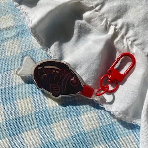 May include: A clear acrylic keychain shaped like a fish-shaped soy sauce packet. The fish has a dark brown body with a red cap at the tail end and a white tail fin. A bright red metal lobster clasp is attached to the red cap of the fish charm.