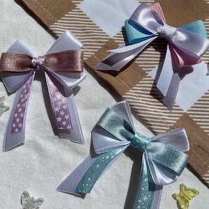 May include: Three handmade satin ribbon bows with white, pink, blue, and teal accents. Each bow has a star pattern on the tails. The bows are arranged on a white fabric background.