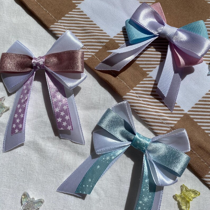 May include: Three handmade satin ribbon bows with white, pink, blue, and teal accents. Each bow has a star pattern on the tails. The bows are arranged on a white fabric background.