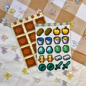 May include: Two sticker sheets featuring pixelated Minecraft items. One sheet reads "item frames" and the other "minecraft items 1". The stickers are brown, yellow, green, blue, and silver. Small, iridescent butterfly decorations are scattered around the stickers.