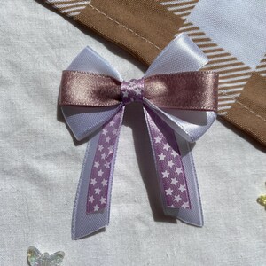 May include: A white and pink satin bow with white stars on the tails. The bow is tied in a simple knot and the tails are long and flowing.