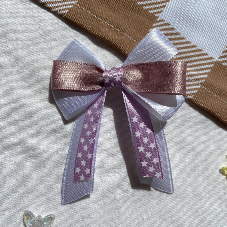 May include: A white and pink satin bow with white stars on the tails. The bow is tied in a simple knot and the tails are long and flowing.