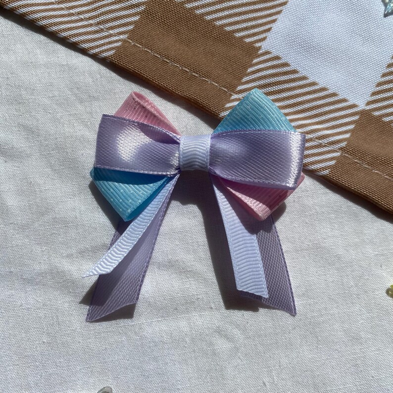 May include: A pastel blue, pink, and white bow with a satin finish. The bow is tied with a white ribbon.