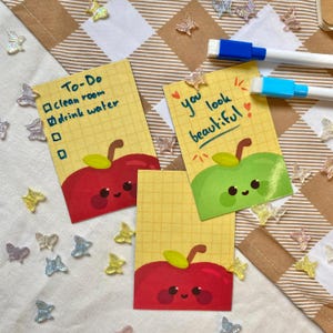 Könnte beinhalten: Drei gelbe Haftnotizen mit Apfelillustrationen und handschriftlichem Text. Eine Notiz sagt "To-Do" mit Kontrollkästchen und Aufgaben. Eine andere sagt "you look beautiful". Dekorative Schmetterlingskonfetti umgibt die Notizen.