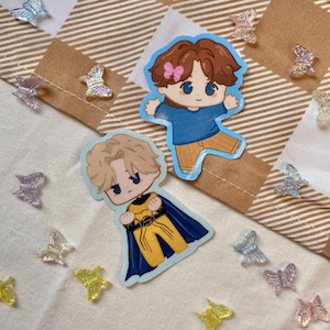 May include: Two cartoon stickers of male characters. One is in a blue top and tan trousers, the other in a yellow and black outfit with a blue cape. Iridescent butterfly-shaped embellishments are scattered around the stickers.