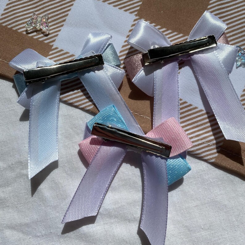 May include: Four pastel-colored satin ribbon hair bows with silver alligator clips. The bows are in shades of light blue, lavender, pink, and a darker blue.