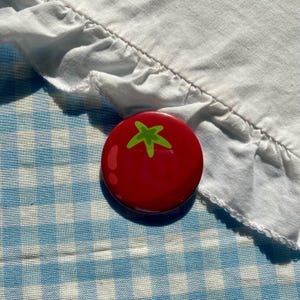 Puede incluir: Una chapa redonda con el gráfico de un tomate rojo brillante y un tallo verde claro. El diseño del tomate incluye un sutil reflejo de luz en el lado izquierdo, lo que crea una apariencia suave y brillante en la superficie del accesorio.