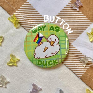 Può includere: Un bottone rotondo con un'anatra dei cartoni animati che tiene una bandiera arcobaleno. Il bottone ha il testo "GAY AS DUCK" su uno sfondo verde con un motivo a quadri. La parola "BUTTON" è scritta verticalmente in alto.