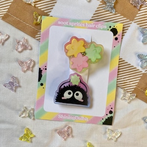 Könnte beinhalten: Zwei Soot Sprite Haarspangen auf einer Karte mit dem Text "soot sprites hair clips" und "ShiningDalbit". Die Spangen zeigen einen schwarzen Soot Sprite und bunte Blumenakzente. Klare, schmetterlingsförmige Dekorationen sind verstreut.