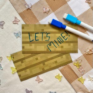 Könnte beinhalten: Zwei braune Karten mit einem pixeligen Holzdesign und dem Text "LET'S MINE!" in Türkis. Zwei weiße Whiteboard-Marker mit blauen Spitzen liegen auf den Karten. Klare, schmetterlingsförmige Dekorationen sind verstreut.