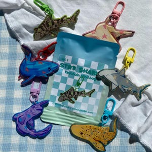 Könnte beinhalten: Eine Sammlung von Hai-förmigen Schlüsselanhängern in verschiedenen Farben und Mustern, darunter Sterne und Punkte. In der Mitte befindet sich ein blauer Beutel mit dem Text "Star Shark Keychain Mind bag". Die Schlüsselanhänger haben unterschiedlich farbige Verschlüsse.