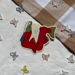 Può includere: Un adesivo di salmone rosso e verde in stile cartone animato. Il salmone sta nuotando con delle bolle intorno a lui. L'adesivo è su uno sfondo bianco.