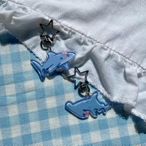 Könnte beinhalten: Zwei Acryl-Anhänger in Form von hellblauen Haien, befestigt an silberfarbenen sternförmigen Verschlüssen. Ein Anhänger zeigt einen Hai mit einer kleinen rosa Schleife, der andere einen Hammerhai mit einem rosa Detail nahe dem Maul.