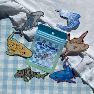 Può includere: Una collezione di adesivi di squali colorati con motivi a stella, tra cui uno squalo martello, uno squalo leopardo e uno squalo blu. È presente anche un sacchetto a quadretti blu e bianco con la scritta "STAR SHARKS".