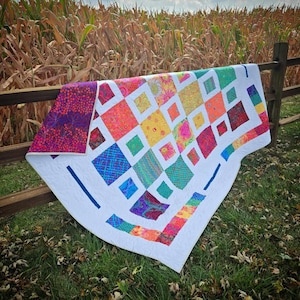 May include: A colorful quilt with a white background and a variety of squares in different colors and patterns. The quilt is hanging on a wooden fence in front of a field of corn.