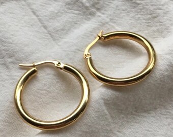 May include: Two gold-colored hoop earrings. The earrings are round and have a shiny finish. They are displayed on a white, textured surface. The earrings are a classic jewelry item.