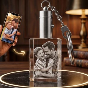 Puede incluir: Un llavero de cristal transparente con una foto de un hombre y un niño. El llavero tiene una parte superior de metal plateado y una cadena con un llavero. Un smartphone muestra la misma foto. El fondo es una superficie de madera oscura.
