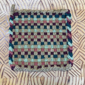 May include: A square, woven potholder in a checkerboard pattern. The potholder features a mix of colors, including teal, purple, pink, and tan. The edges are braided with teal and purple yarn, and a small loop is attached for hanging.