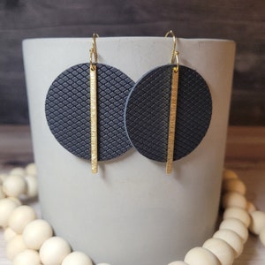May include: A pair of dark gray, textured leather earrings with a gold-colored vertical bar. The earrings are circular and hang from gold-colored hooks. The earrings are displayed in front of a light gray ceramic container.