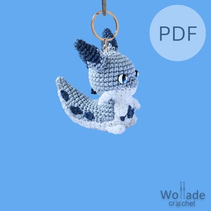 Puede incluir: Un llavero de amigurumi de crochet azul y blanco de un personaje de Pokemon, Eeveelution, con un llavero dorado. El llavero está colgado sobre un fondo azul claro. El texto "PDF" es visible en un círculo blanco en el lado derecho de la imagen.